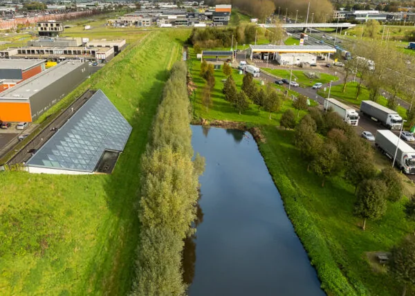 Luchtfoto van het Mart Stamhof met een driehoekig glazen gebouw, een watergang, omliggend groen en een nabijgelegen snelweg met verkeer.